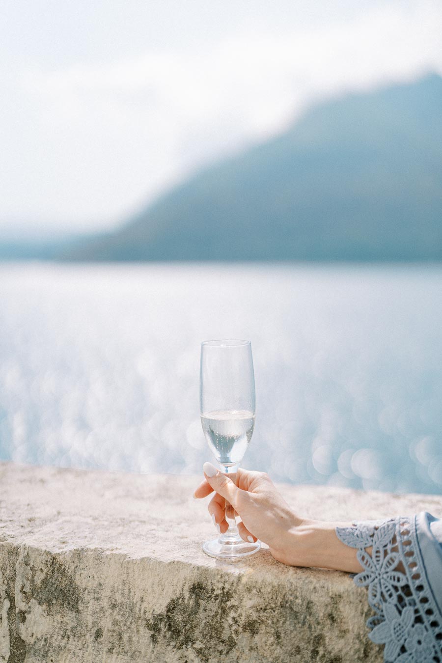 Hand holding a champagne flute with a scenic lake and mountain view in the background, capturing a serene and elegant moment by the water.
