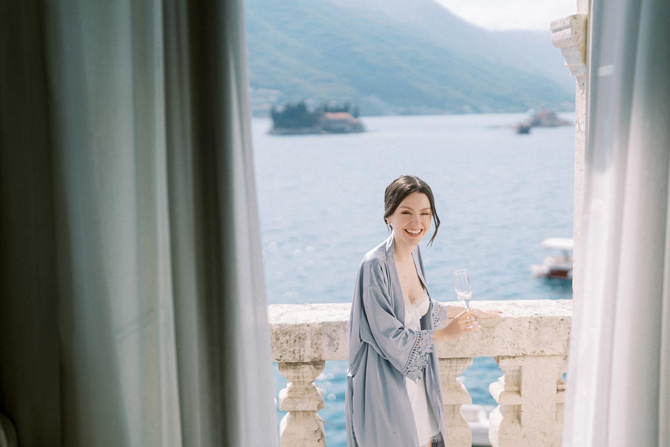 Woman in blue robe smiling while holding a champagne glass on a balcony overlooking a scenic ocean view.