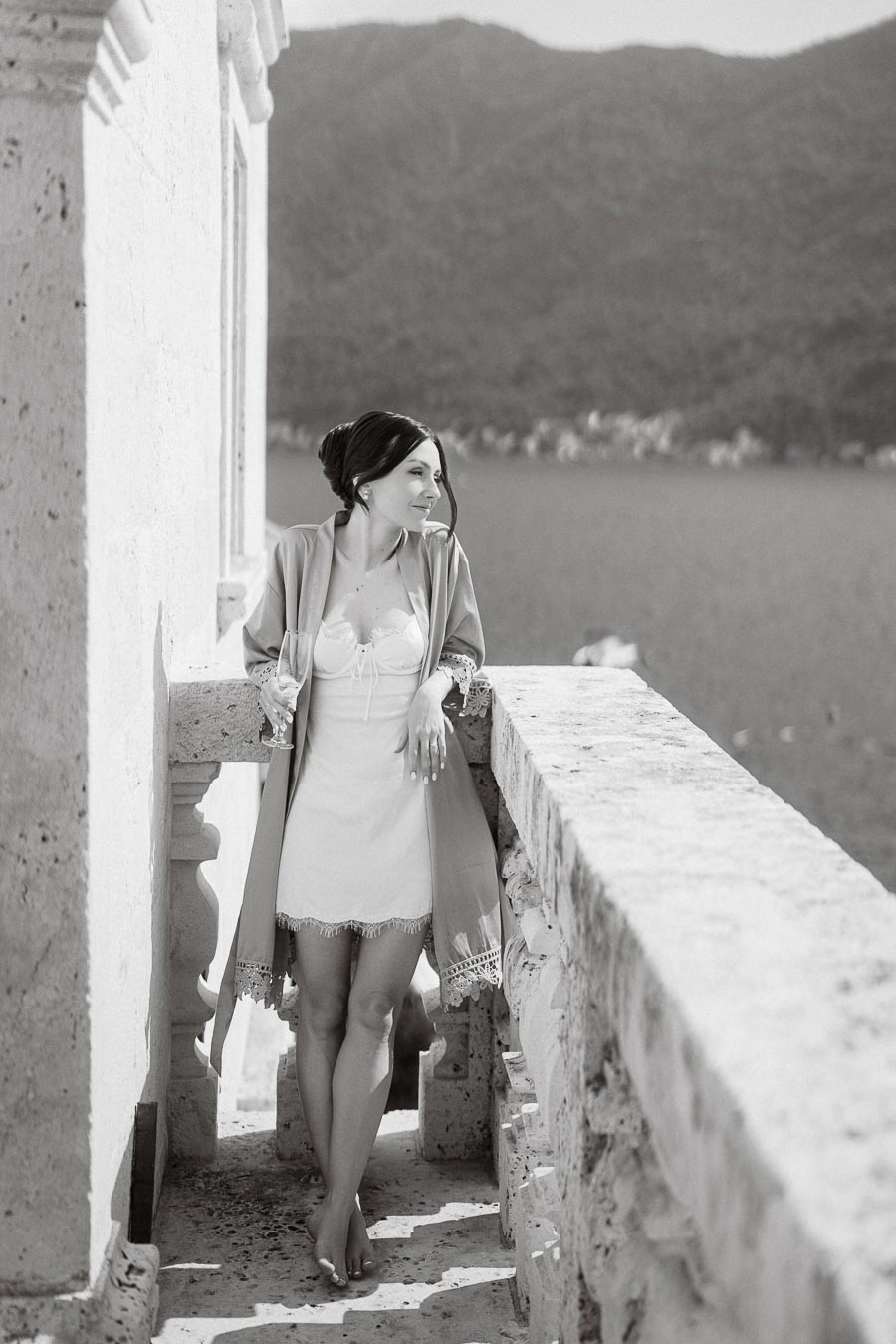 Black and white photo of a woman in a lightweight dress and robe, holding a champagne glass, standing on a stone balcony overlooking a serene lake and distant mountains.
