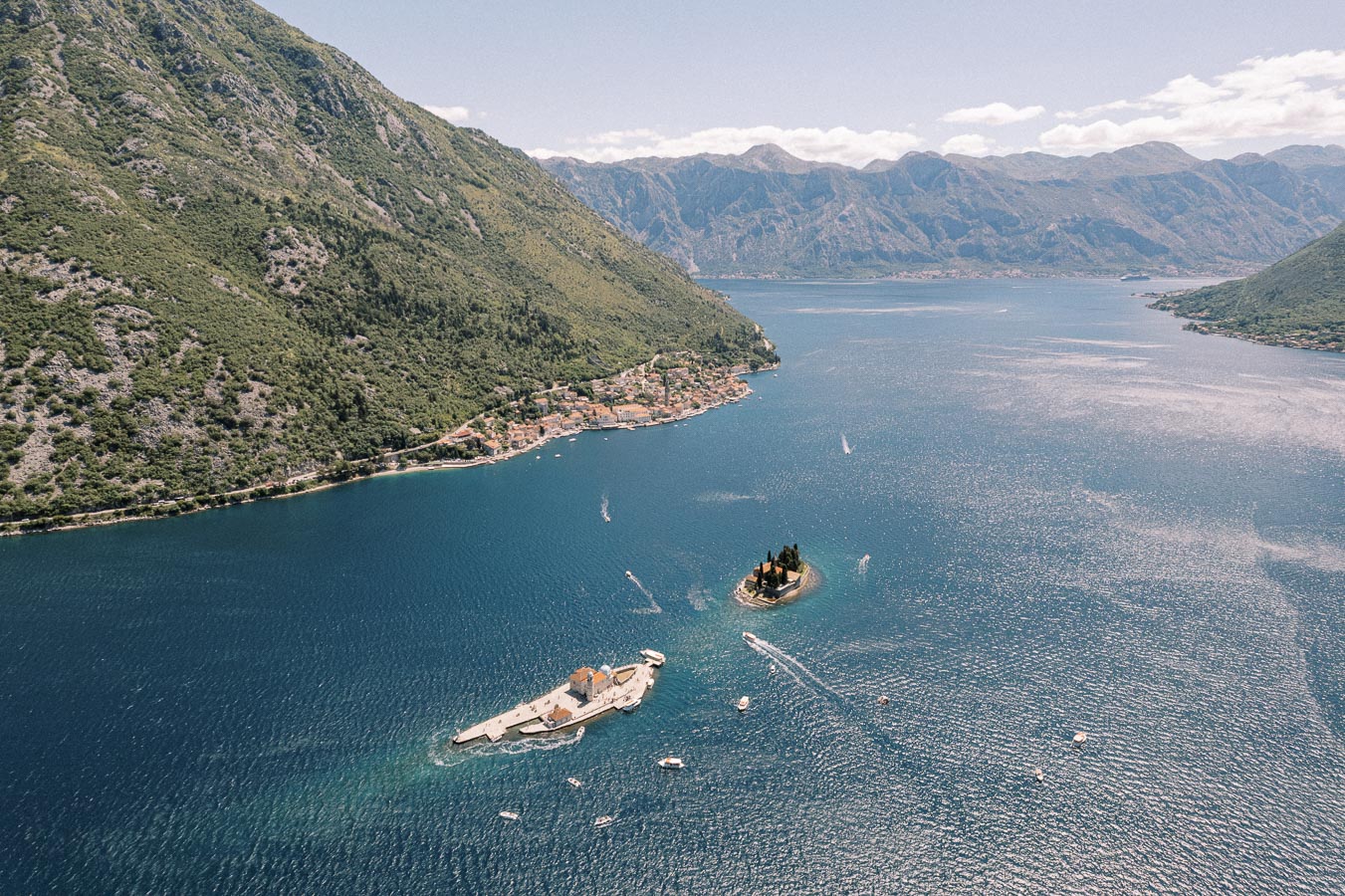 Aerial view of the Bay of Kotor in Montenegro, showcasing a small coastal town nestled by the lush green mountains, two picturesque islands with historic buildings, and several boats navigating the serene blue waters.