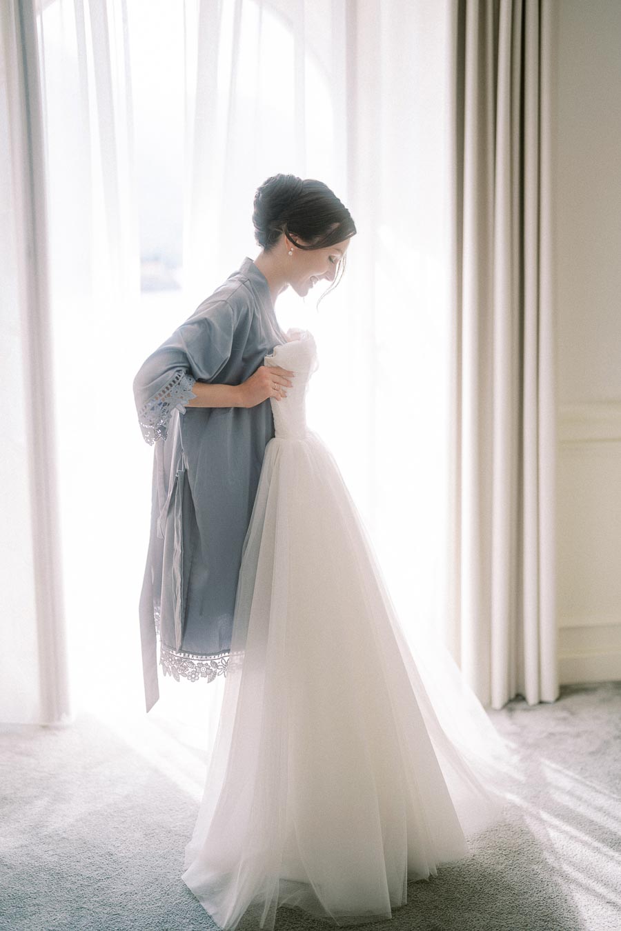 Bride in light blue robe admiring her elegant white wedding dress by a sunlit window.