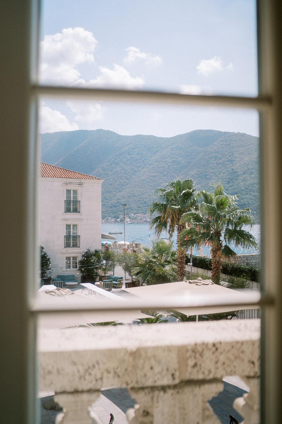 Scenic view of a Mediterranean coastline with mountains, palm trees, and a charming white building, captured through an open window frame.