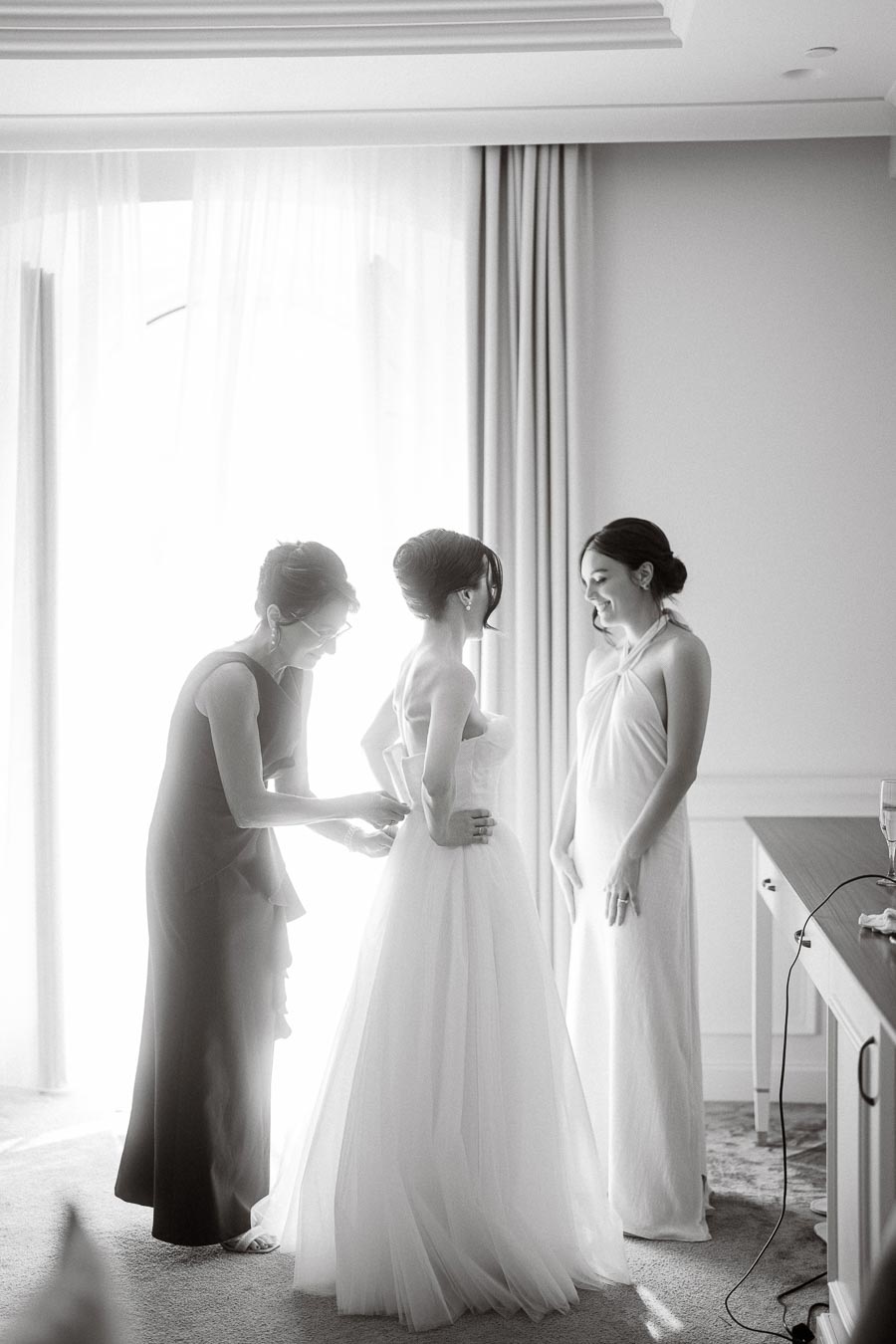 Black and white image of a bride getting ready, assisted by two women in elegant dresses, in a sunlit room with curtains in the background.