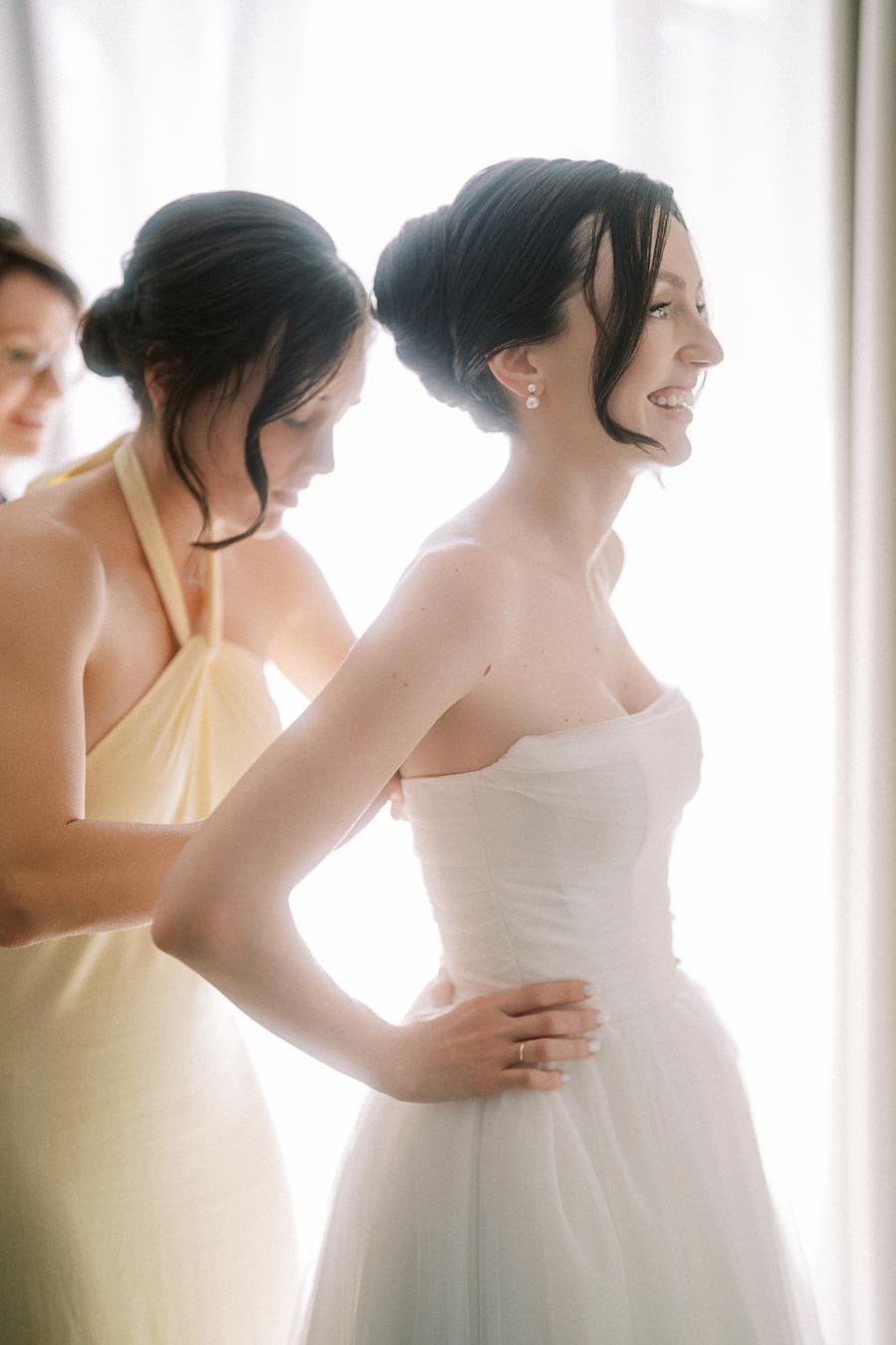 A bride smiles as a bridesmaid helps her with her dress on her wedding day, with soft natural light illuminating the scene.