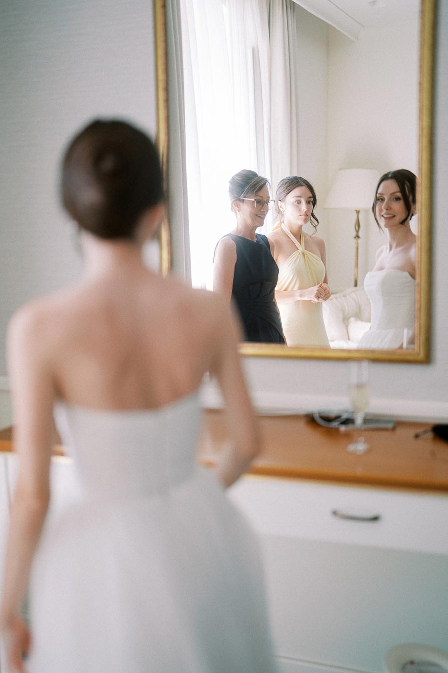 Bride and bridesmaids getting ready for a wedding, reflected in a mirror with soft natural light.