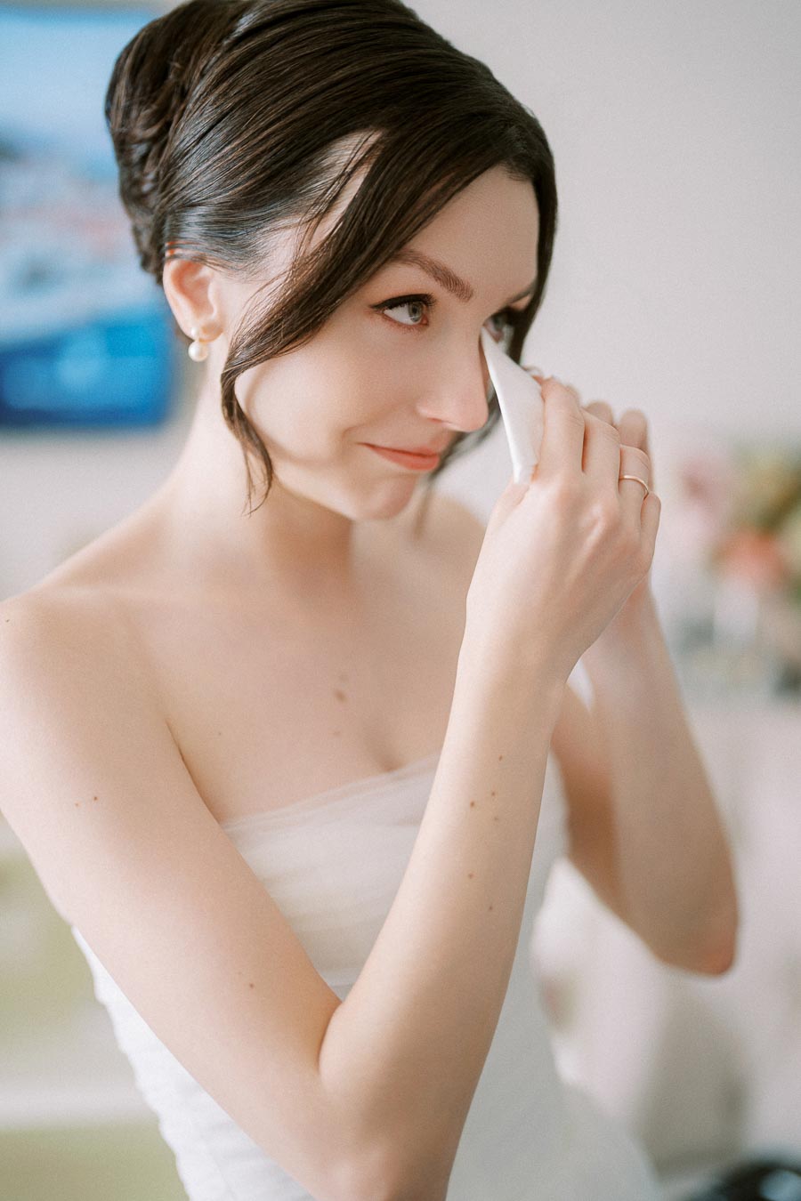 A bride in a strapless wedding dress wipes her tears with a tissue, showcasing an emotional moment before her wedding ceremony.