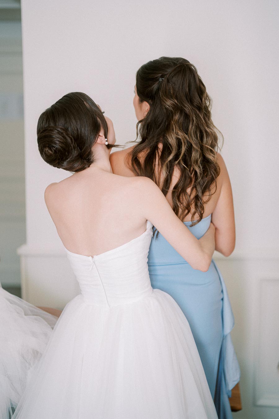 Two women embracing, viewed from behind. One is wearing a white wedding dress, and the other is in a light blue gown.