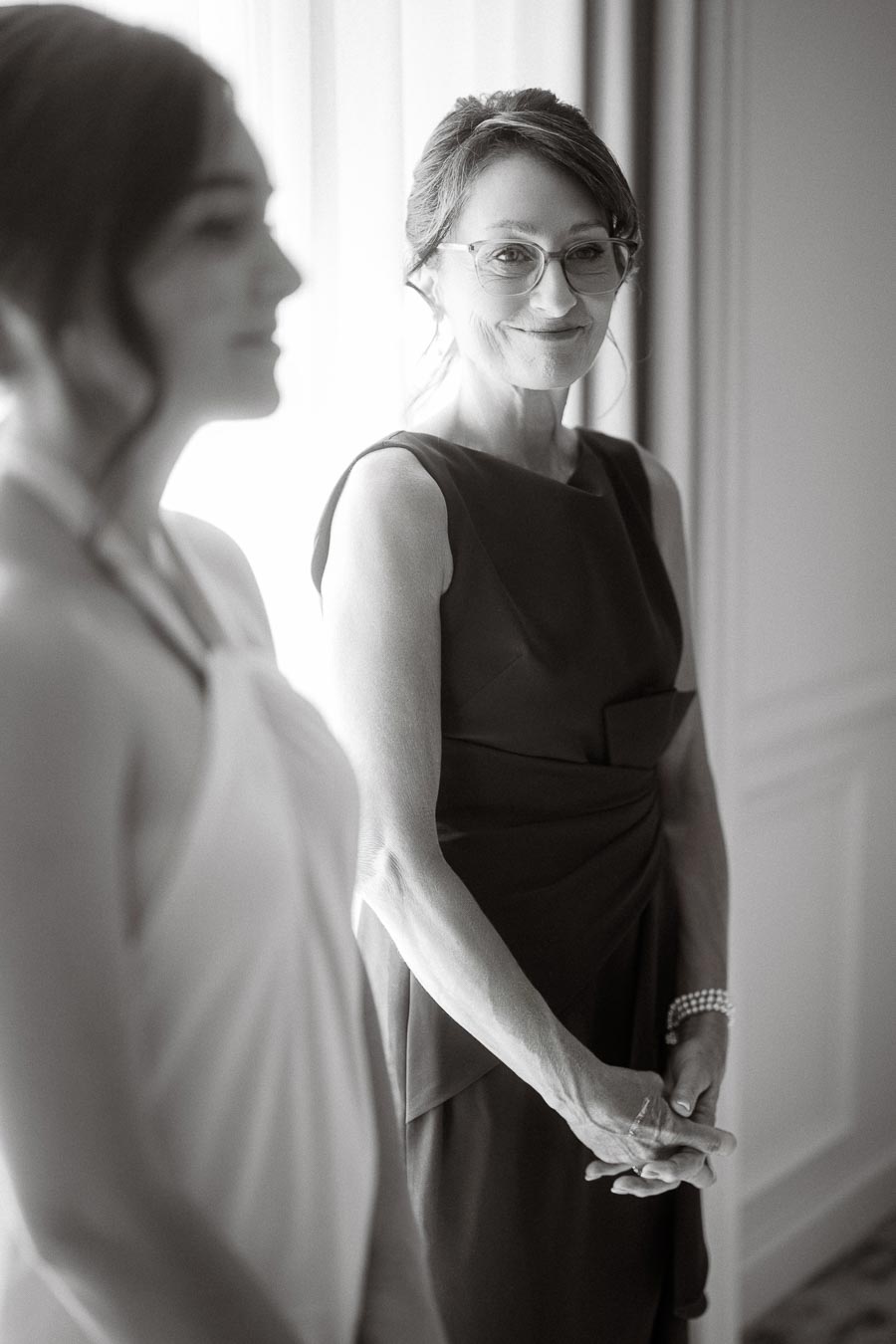 A grayscale portrait features two women in elegant attire, standing near a window. The woman in focus smiles softly, wearing glasses and a sleeveless dress, showcasing a calm and poised demeanor in a serene setting.