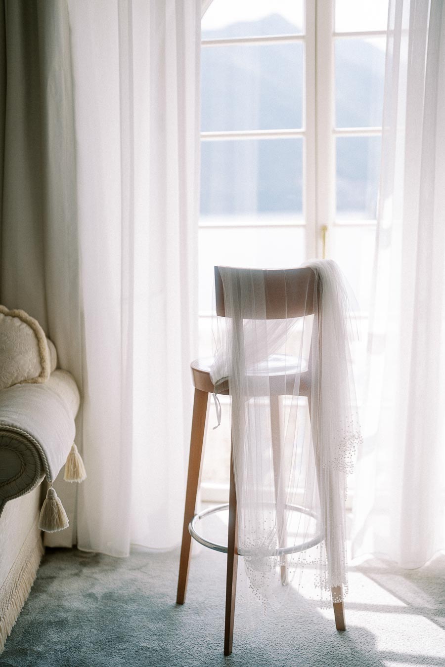 Elegant bridal veil draped over a wooden chair in a sunlit room with soft curtains and a picturesque window view.