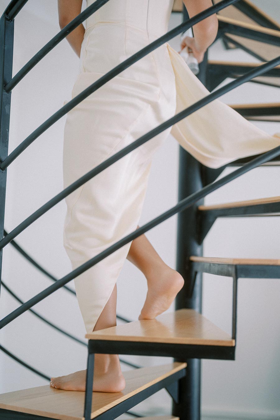 A person in an elegant cream dress walking up a modern spiral staircase barefoot, highlighting grace and contemporary design.