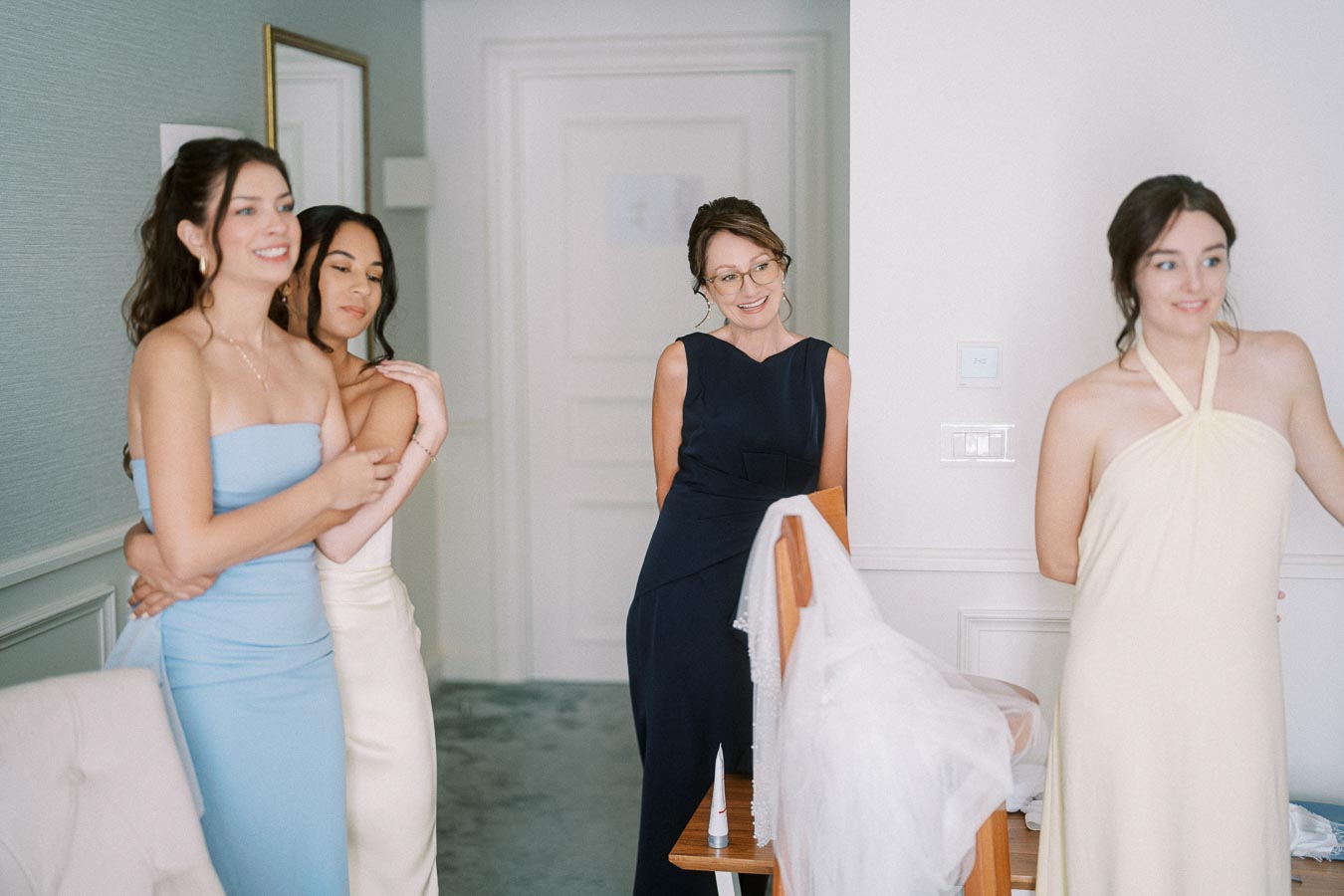 Four women in elegant dresses are gathered in a brightly lit room, smiling and enjoying a moment together. The atmosphere appears joyful and celebratory, possibly suggesting a special occasion or gathering.