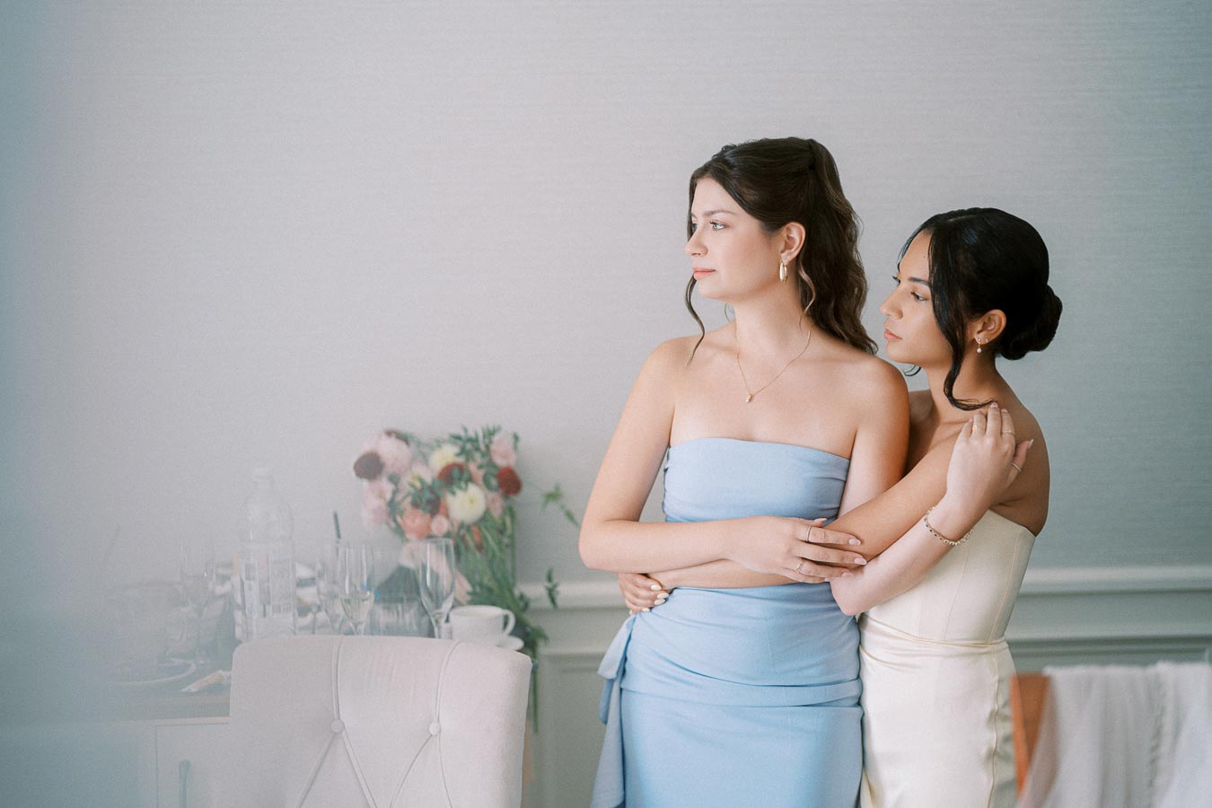 Two women in elegant strapless dresses, one in light blue and the other in white, embrace each other in a softly lit room. A table with a bouquet of flowers and glassware is blurred in the background.