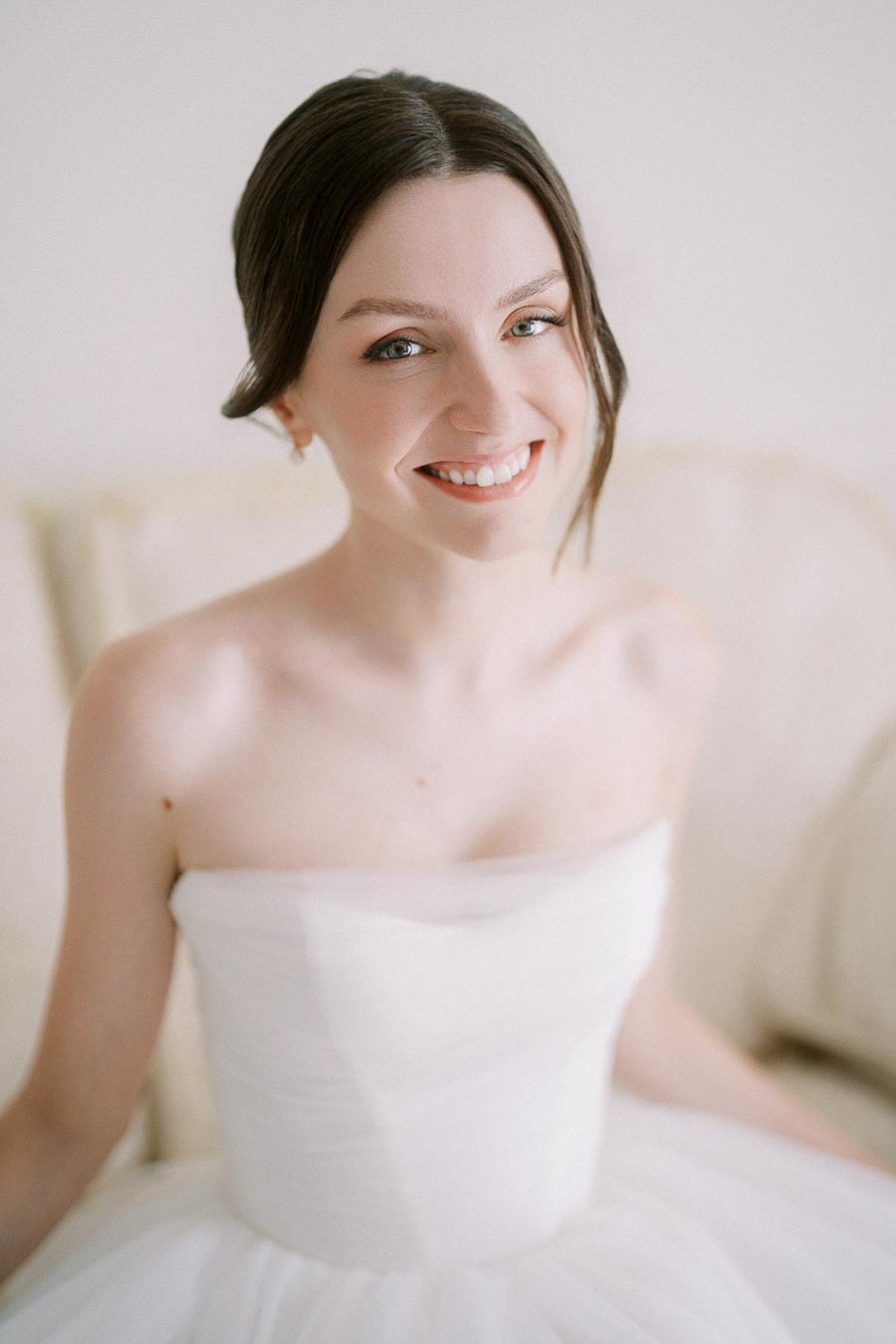 A bride smiling in a strapless white wedding dress, with natural makeup and dark hair styled in an elegant updo, seated on a light-colored sofa, exuding joy and elegance.