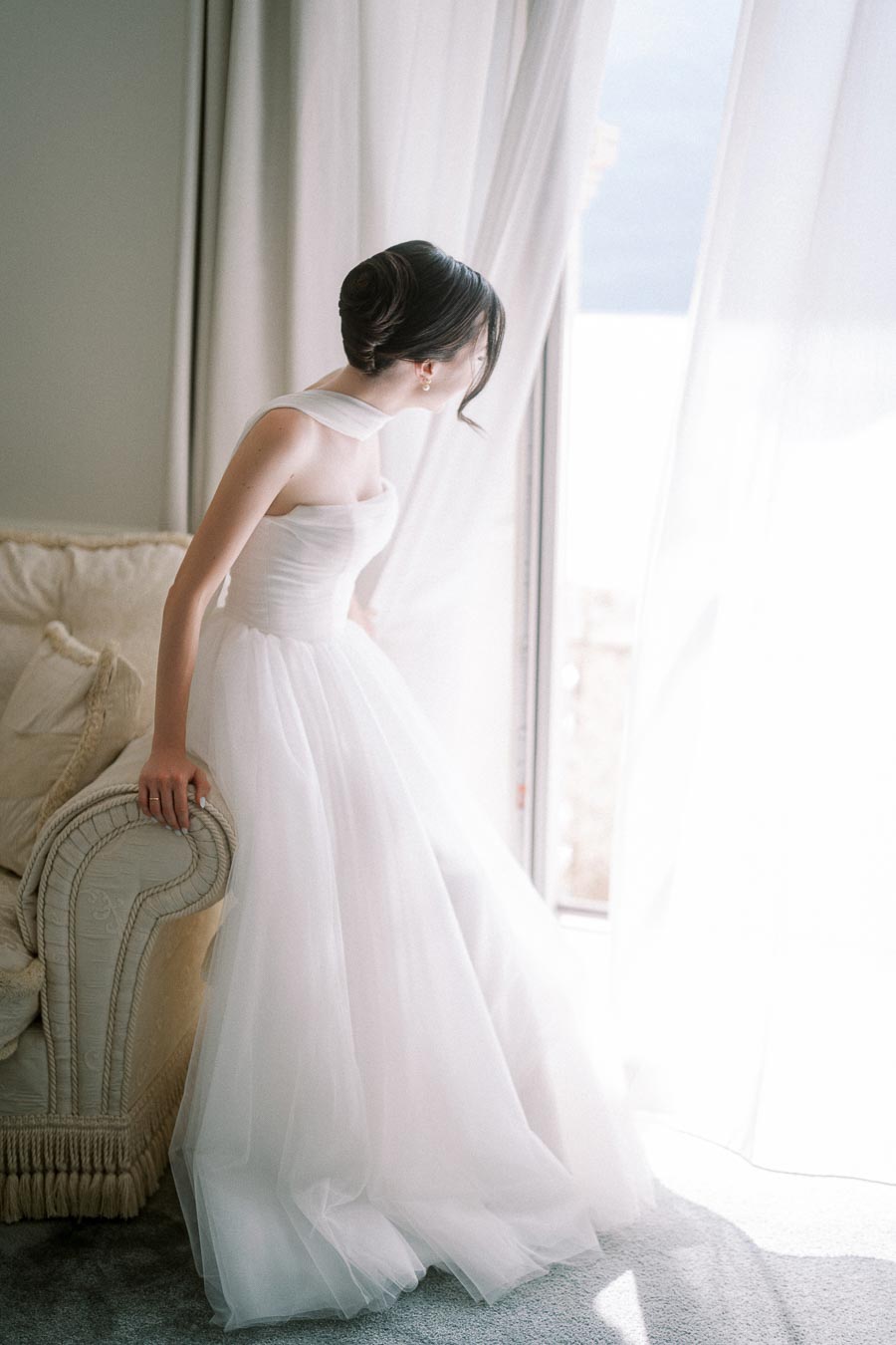 Elegant bride in a white wedding dress gazing out the window with soft natural light, creating a serene and romantic atmosphere.