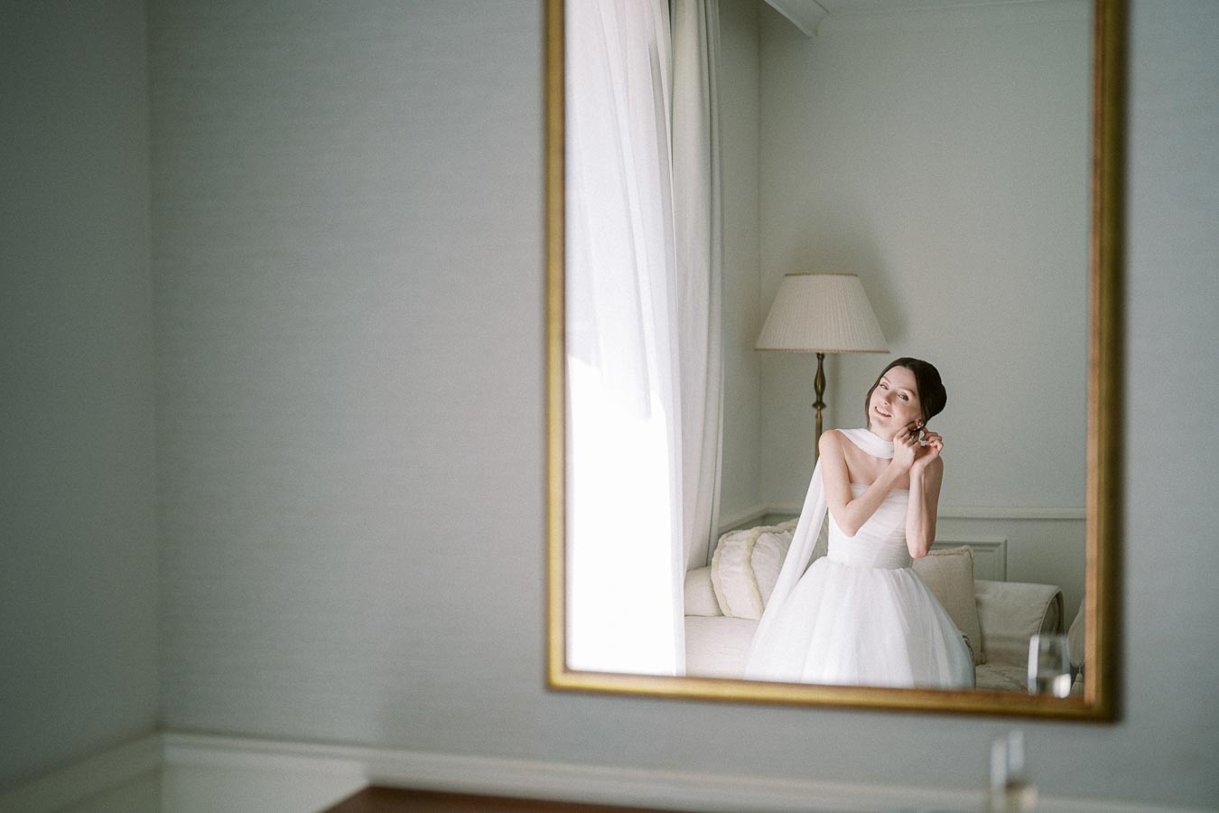 Bride in a white gown adjusts her earring, reflected in a large mirror, with soft lighting in an elegant room setting.