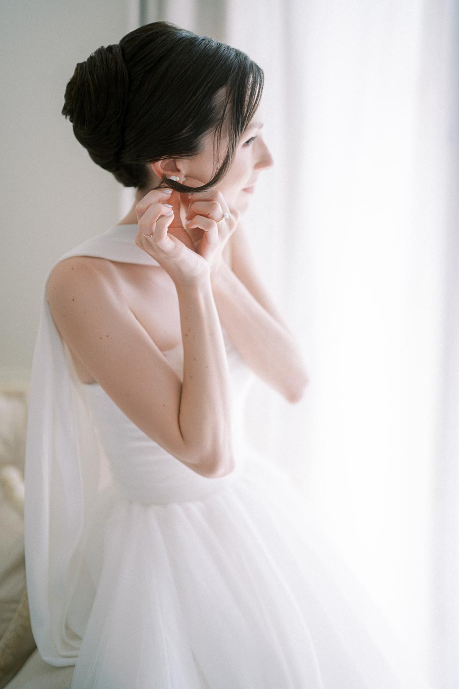 Elegant bride adjusting earrings while preparing for wedding in soft natural lighting, wearing a white gown.