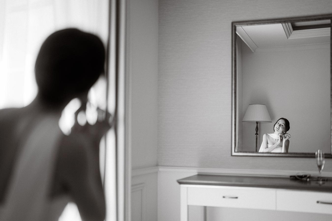 Black and white image of a woman seen in a mirror, elegantly adjusting her earring. The mirror reflection shows her in a well-lit room with a floor lamp, creating a soft and serene atmosphere.