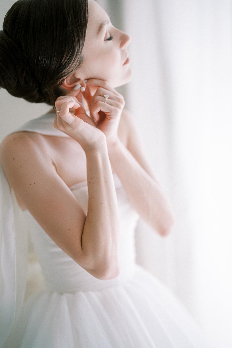 A bride gently adjusts her pearl earring, her hair elegantly styled in an updo, wearing a delicate white wedding gown in a softly lit room.