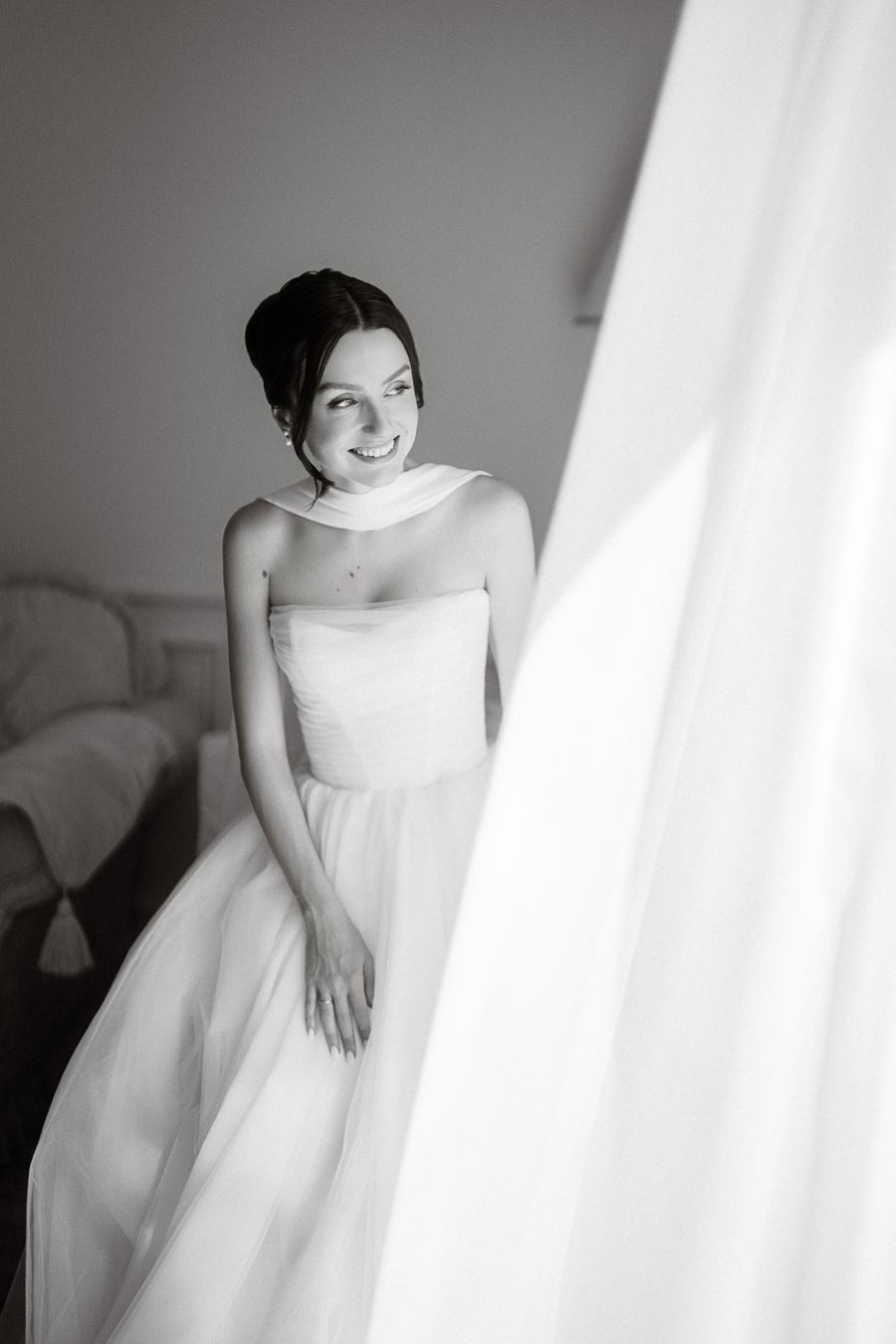 Black and white photo of a smiling bride in an elegant strapless wedding dress, looking out a window, in a softly lit room.