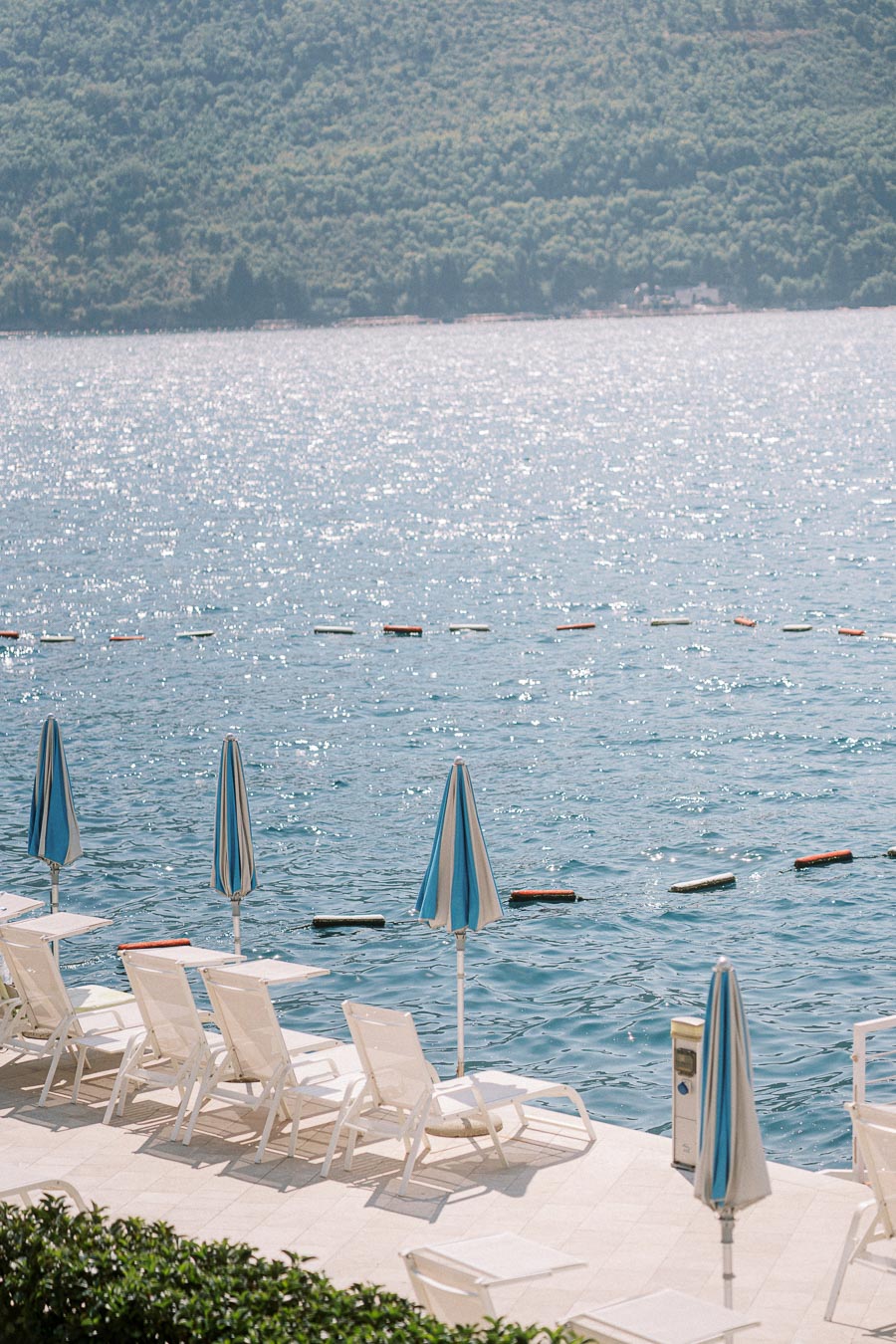 Peaceful lakeside view with empty lounge chairs and blue-striped umbrellas on a sunlit patio, sparkling water and lush green hills in the background, ideal summer relaxation spot.