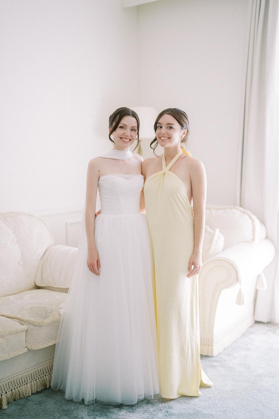 Two women smiling in elegant dresses, one in a white wedding gown and the other in a light yellow halter gown, standing together in a softly lit room with a cream sofa.