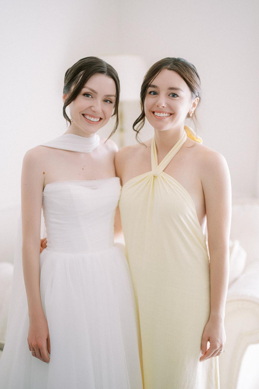 Two smiling women dressed in elegant formal attire, one wearing a strapless white dress and the other wearing a pale yellow halter neck dress, posing closely in a softly lit room, capturing a moment of happiness and friendship.