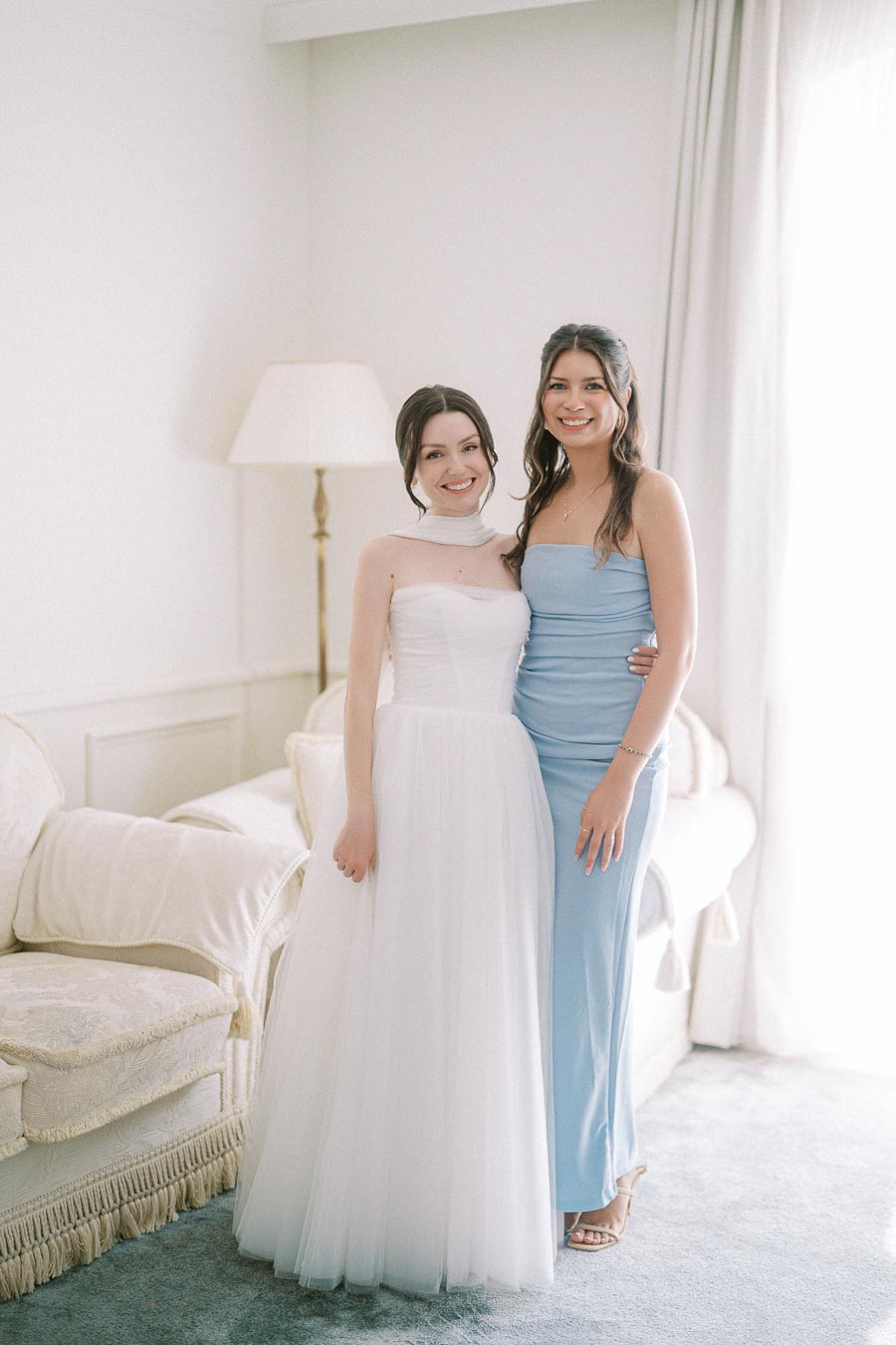 Two women posing together in a bright room; one wearing a white formal dress and the other in a light blue gown, both smiling warmly, next to a plush couch and lamp.