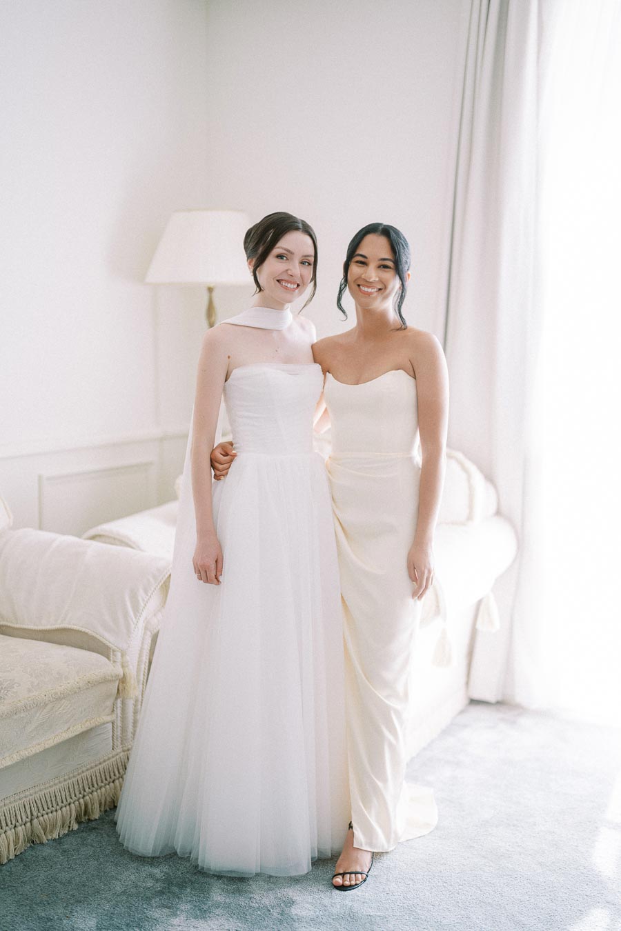Two women in elegantly styled white dresses posing together in a softly lit room, ideal for capturing the essence of a serene wedding atmosphere.