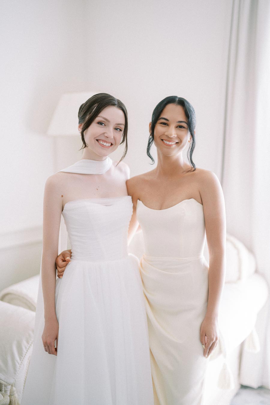 Two women in elegant white dresses smiling and posing together in a softly lit room.