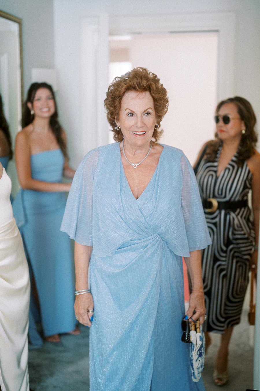 Elegant older woman in a light blue dress smiling, surrounded by people in stylish attire, creating a joyful and celebratory atmosphere indoors.