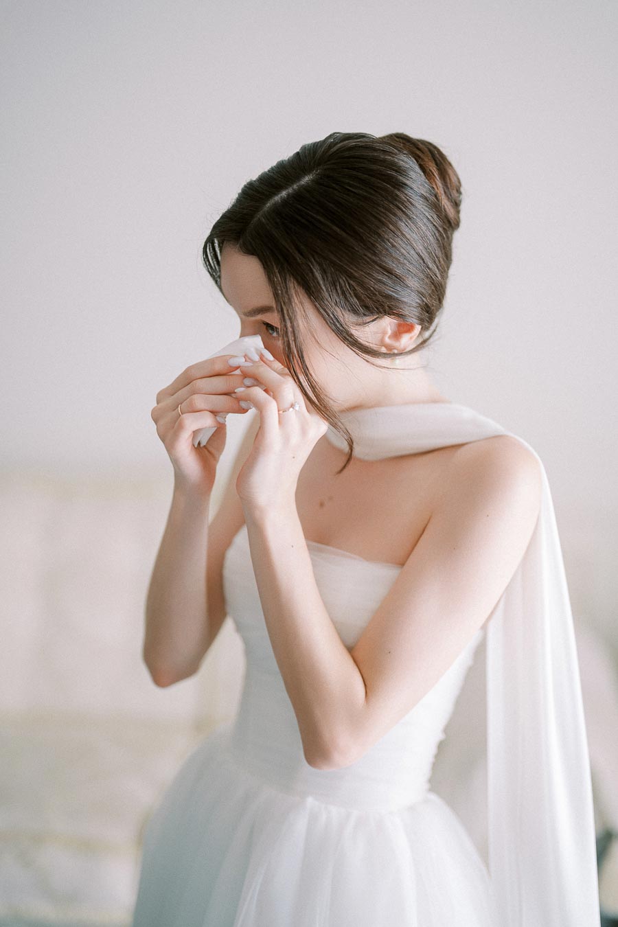 A bride in a white wedding dress gently dabbing her face with a tissue, showcasing an elegant updo and soft natural lighting.
