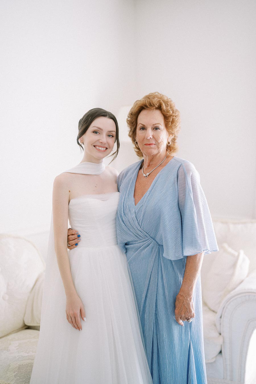 A bride in a white dress smiling with an older woman in a light blue gown, standing indoors in a softly lit room.
