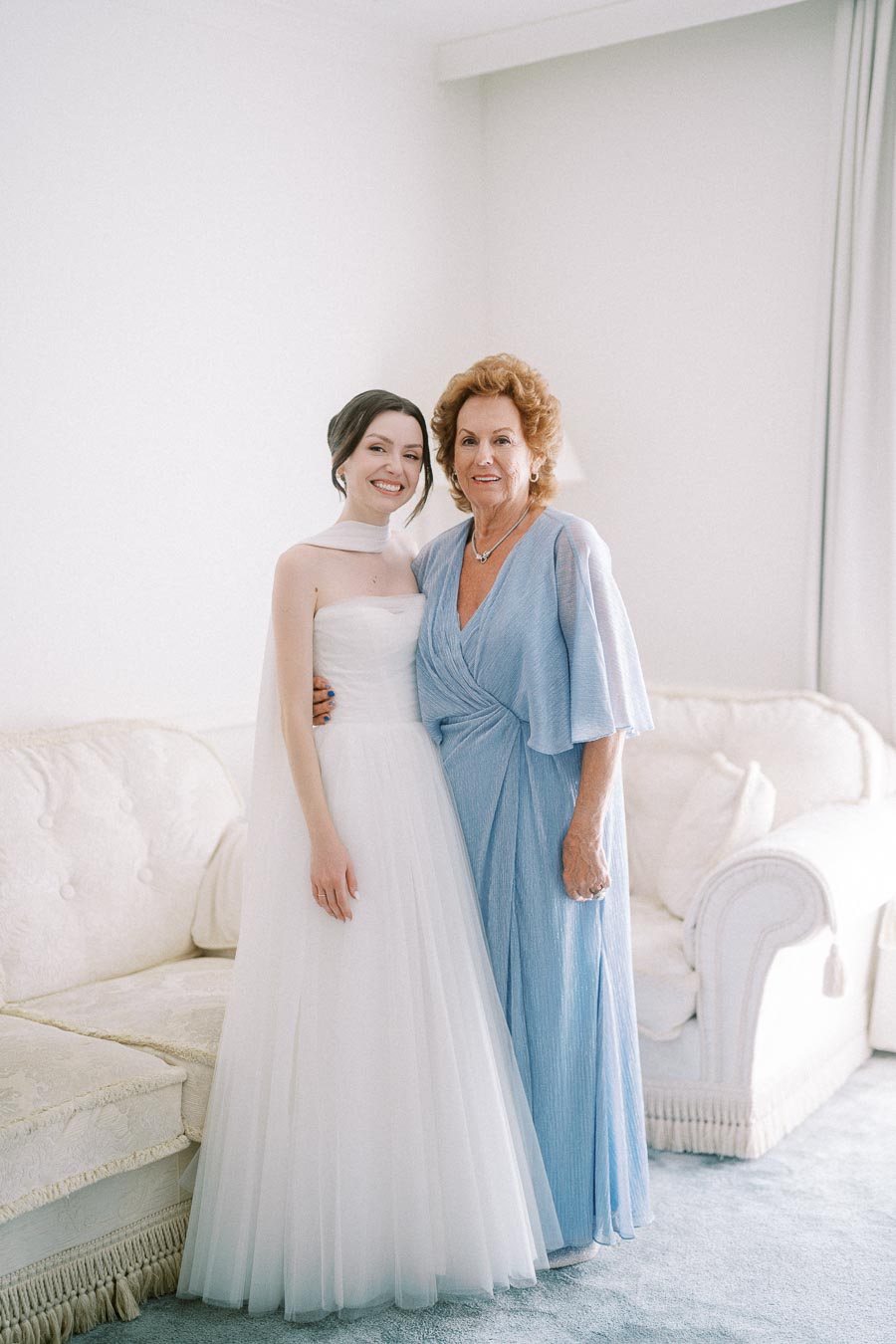 A bride in a white wedding gown stands smiling next to an older woman in a light blue dress, posing in a bright, elegant room with a white sofa.