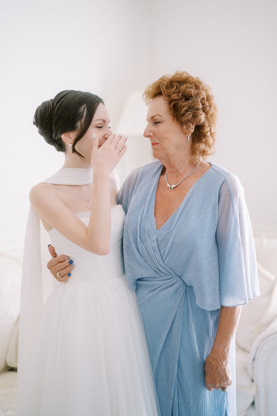 Young bride in white dress shares an emotional moment with elderly woman in a blue dress, capturing a tender pre-wedding scene indoors.