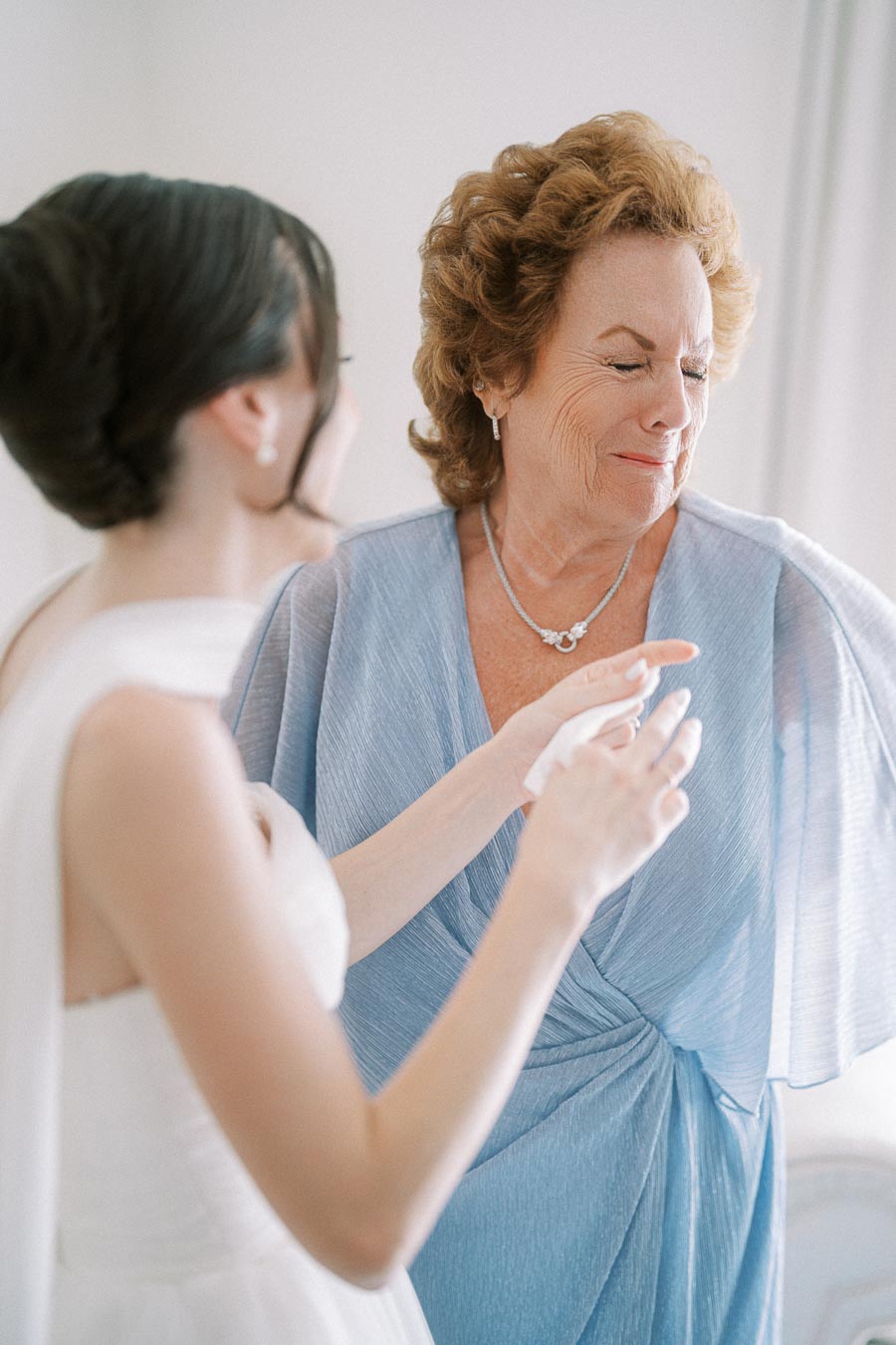 Senior woman in a light blue dress sharing an emotional moment with a bride in a white gown, both smiling warmly.