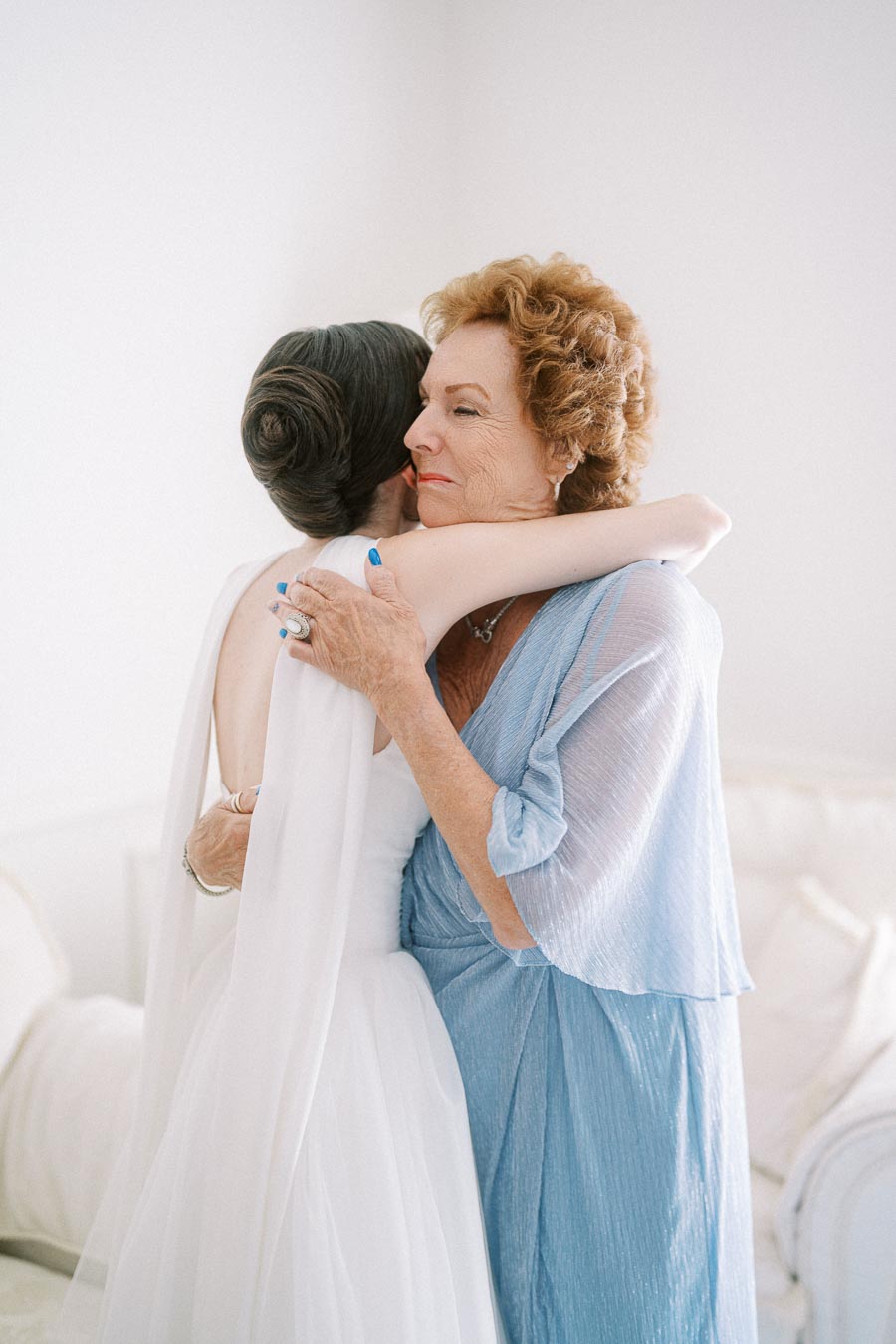 Elderly woman in blue hugging a bride in a white gown, sharing a warm embrace in a softly lit room.