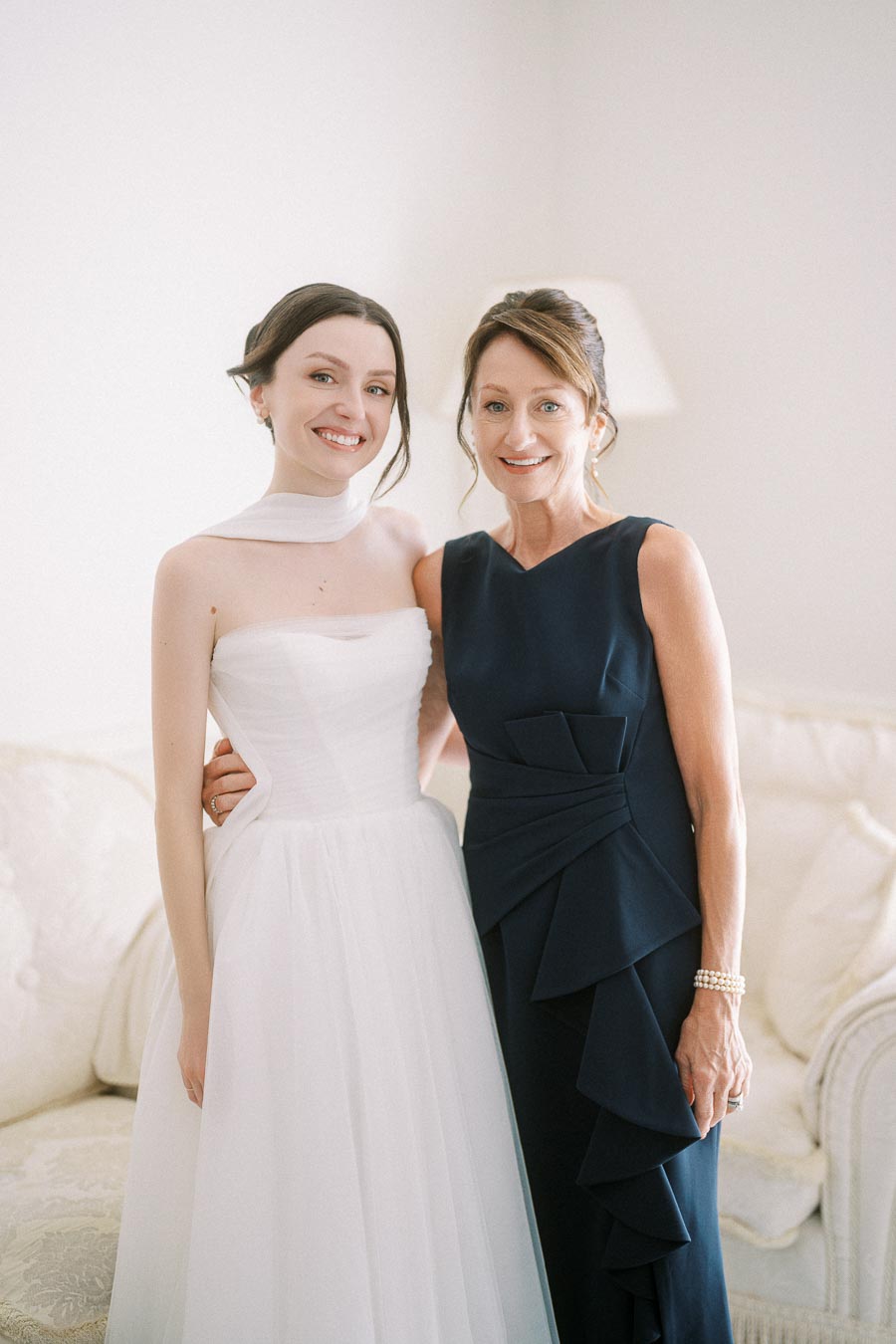 Smiling bride in elegant white wedding dress standing with woman in stylish navy gown, both posing indoors.