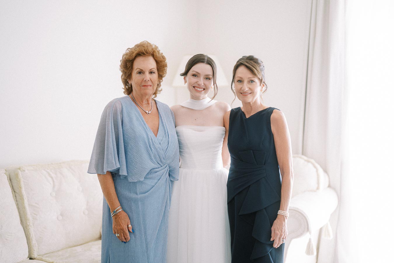 Three women posing together in elegant dresses, standing in a well-lit room with a light sofa in the background, exuding a warm and joyful atmosphere.
