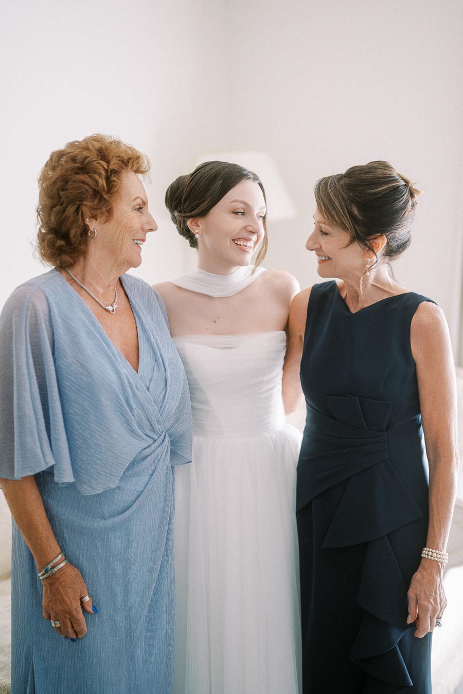 Three women smiling together, with one in a bridal gown and the others in elegant dresses, sharing a joyful moment. Perfect for wedding and family celebration themes.