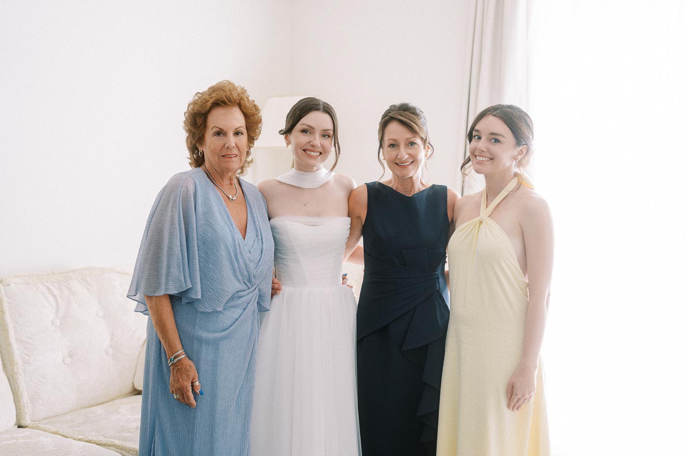 Four women posing together in elegant dresses, including a bride in a white gown, surrounded by family in pastel and dark colors, creating a joyful and intimate wedding atmosphere.