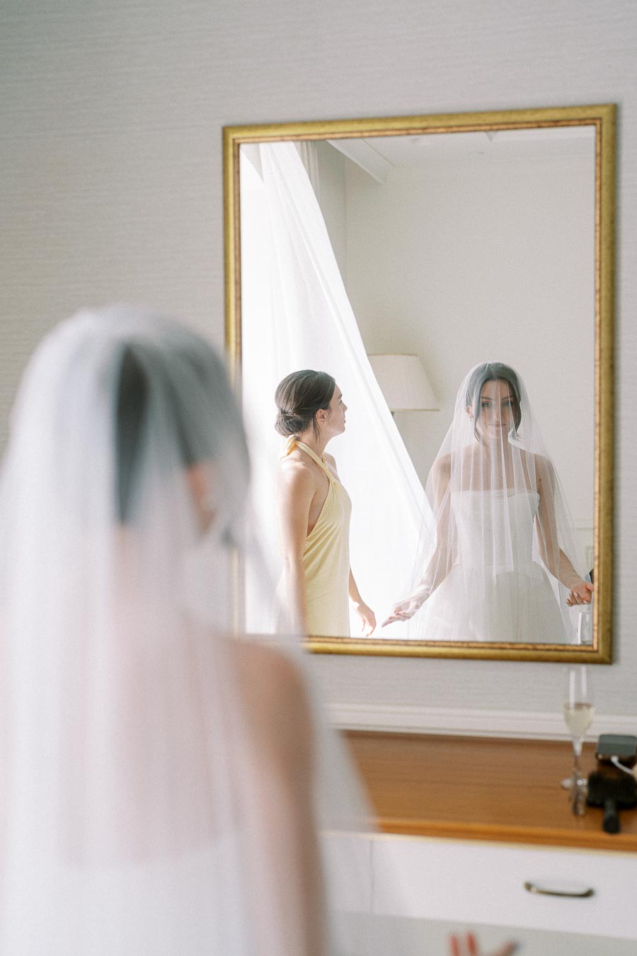 Bride in white wedding dress and veil stands in front of a mirror, with a bridesmaid in a yellow dress adjusting the veil, creating a serene and elegant wedding preparation scene.