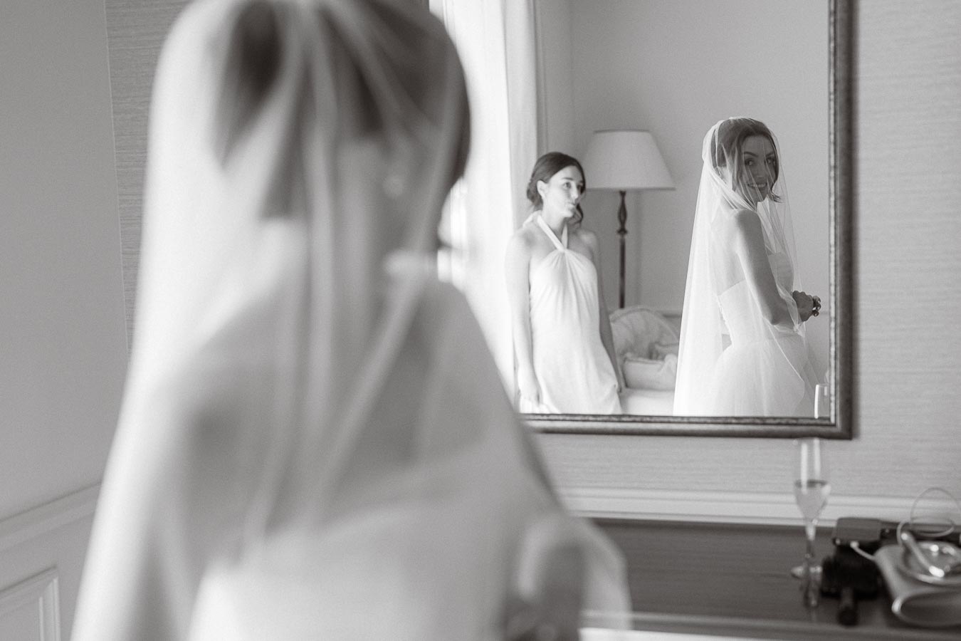 A bride in a white wedding dress and veil smiles at her reflection in the mirror while a bridesmaid stands nearby, creating an elegant and intimate pre-wedding moment.