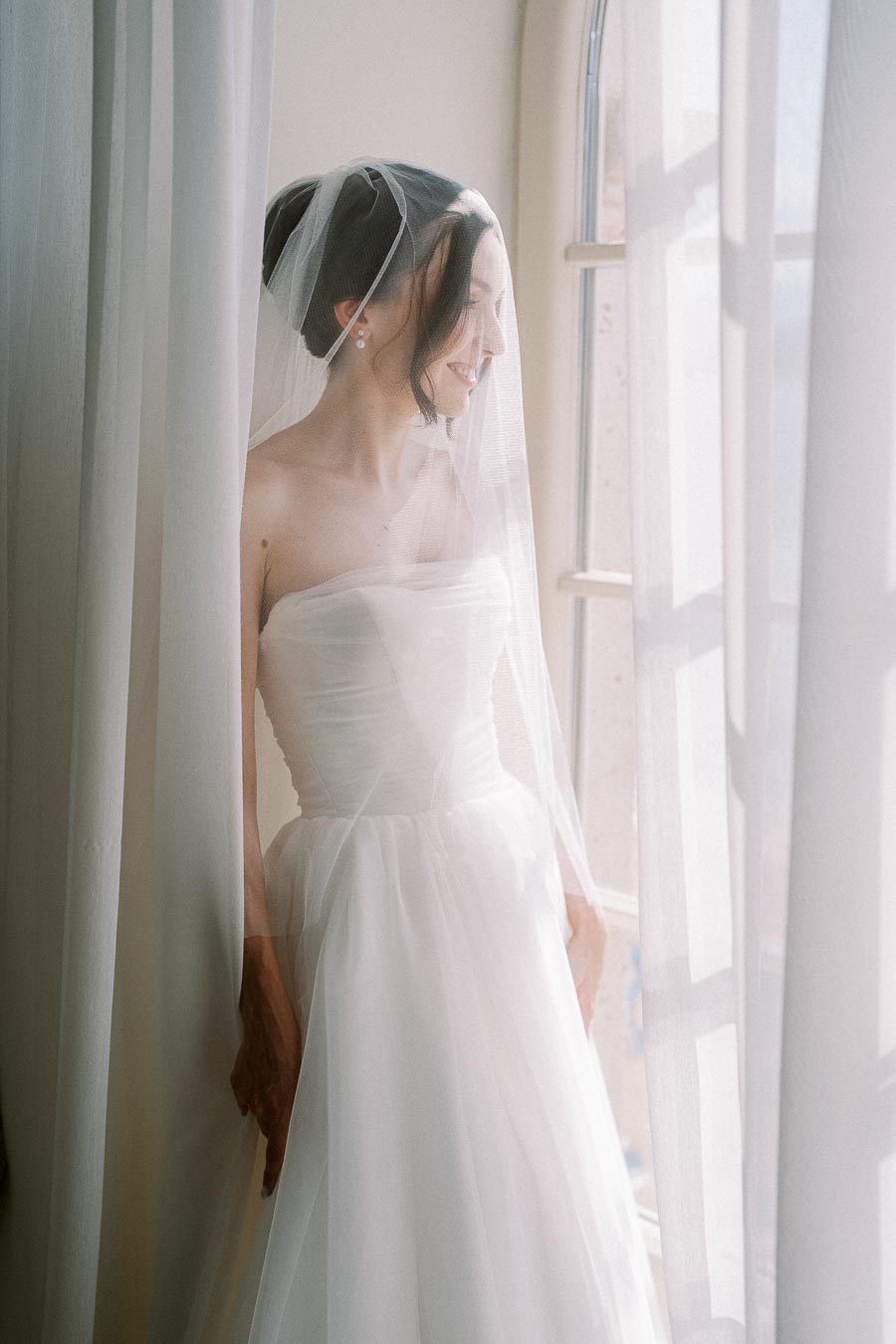 Bride in elegant white wedding dress and veil standing by a window, looking outside with soft natural light filtering through sheer curtains.