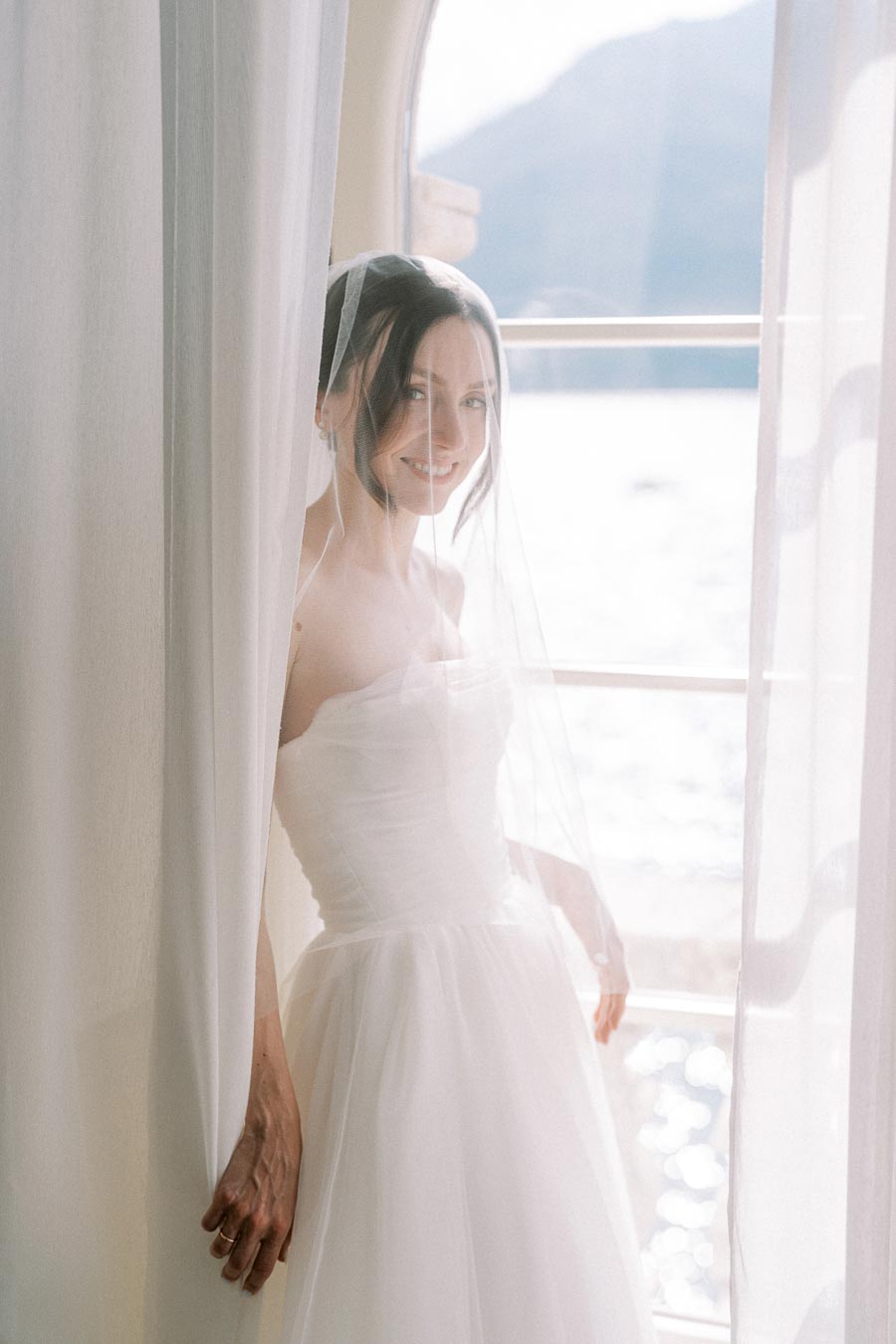 A bride in a white wedding dress smiling while standing by a window with sheer curtains, soft light enhancing the serene ambiance.