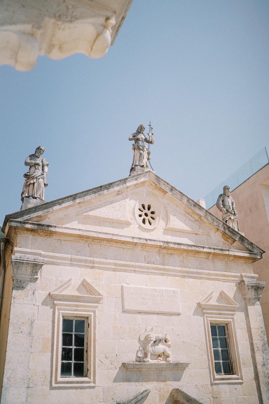 Historic stone building facade with statues under a clear blue sky, featuring intricate architectural details and ancient sculptures on the rooftop.