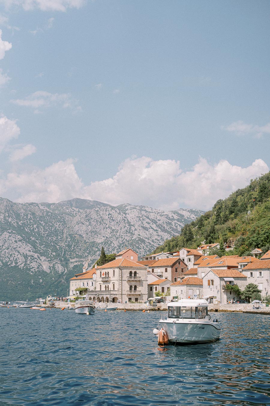 Coastal village scene with rugged mountains in the background, a clear blue sky, and a boat floating on the serene water near charming stone houses with red rooftops. Perfect travel destination for a tranquil escape.