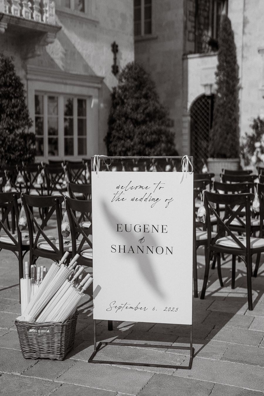 Elegant black and white wedding welcome sign reading 'Welcome to the wedding of Eugene & Shannon, September 6, 2025' set against a charming outdoor venue with wooden chairs and a basket of umbrellas.