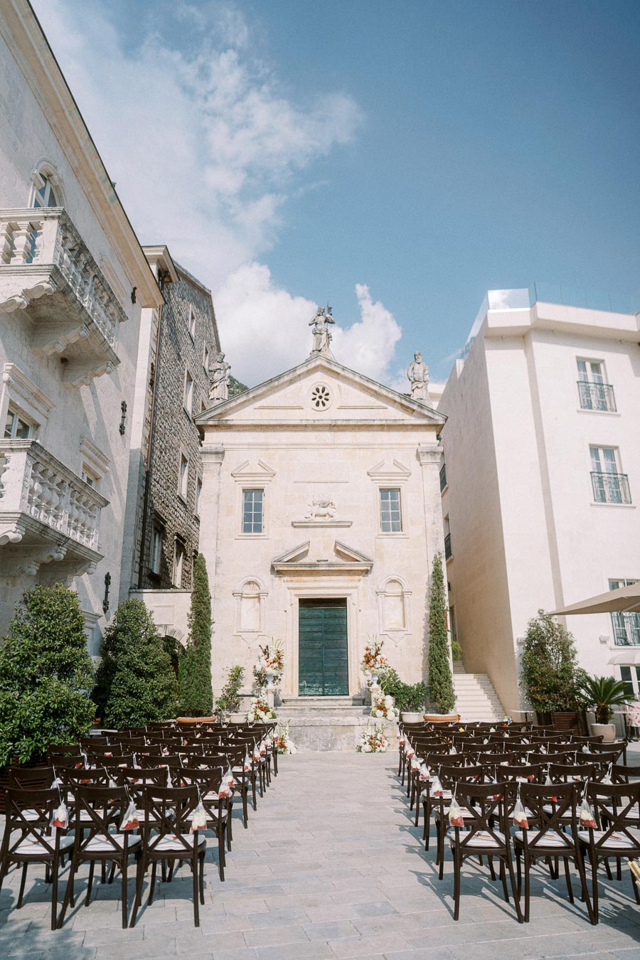 Outdoor wedding ceremony setup in front of a historic stone church with rows of chairs and floral decorations under a clear blue sky.