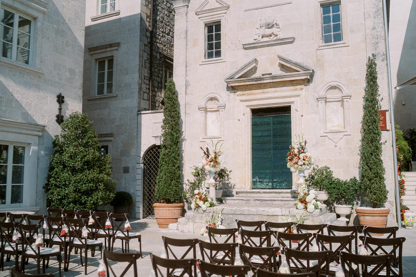 Elegant outdoor wedding ceremony setup with wooden chairs and floral decorations in front of a rustic stone building.
