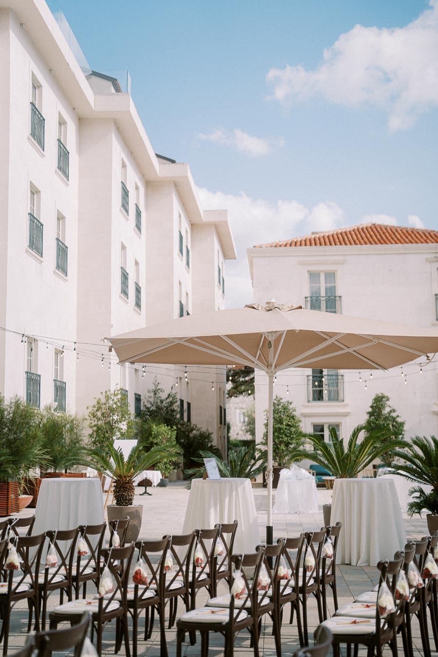 Outdoor wedding venue setup with elegant chairs arranged for guests, large patio umbrella providing shade, and nearby lush greenery, set against a backdrop of modern white buildings and a clear blue sky.